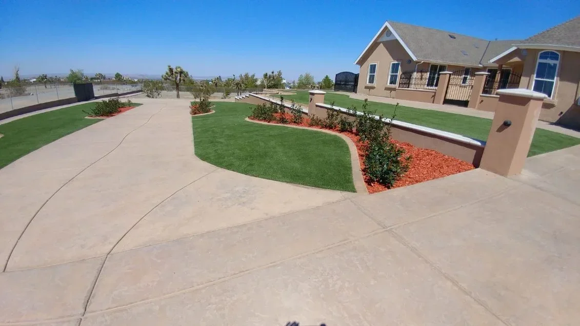 Synthetic turf lawn with red rock mulch beds, curved concrete walkways, and landscape edging