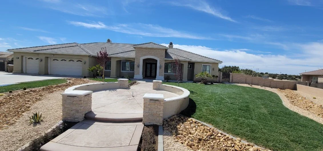 Curved concrete patio with stone pillars, sod lawn, and river rock landscaping
