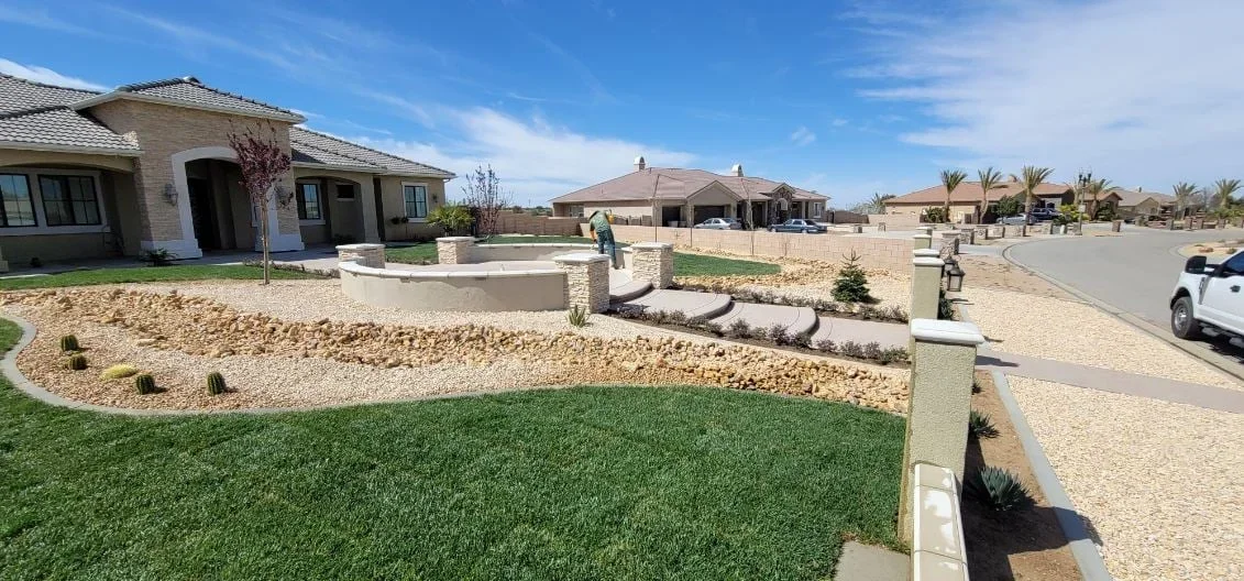 Front yard with synthetic turf, rock beds, desert plants, concrete curbing, and stone pillars