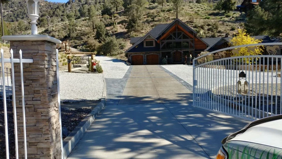 Concrete driveway with stone pillars, white gate, and gravel borders leading to mountain home