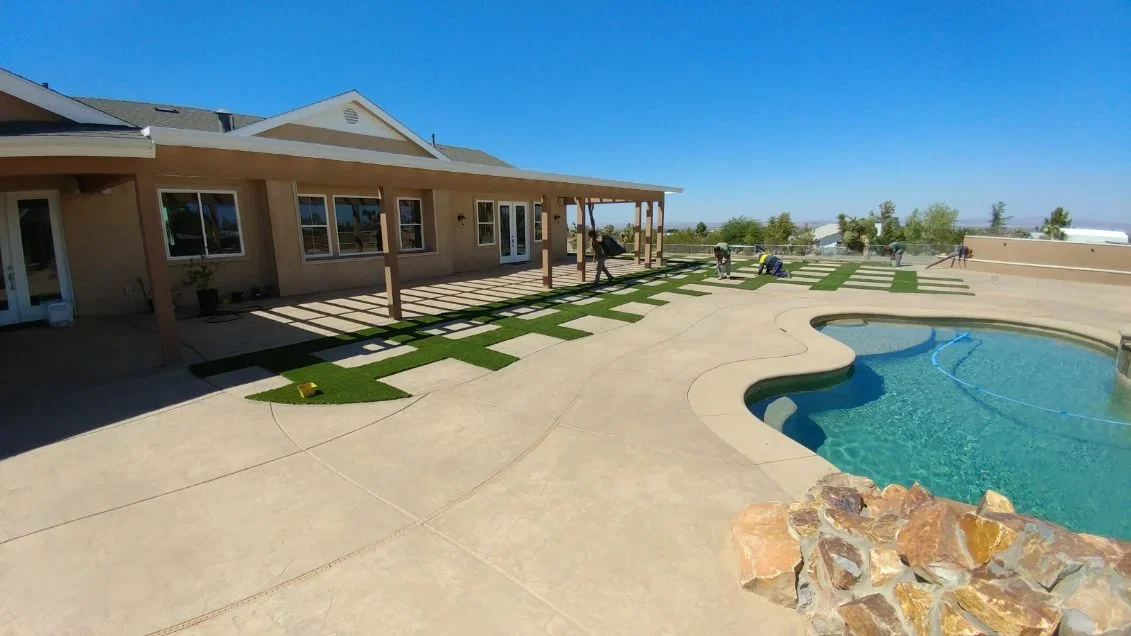 Pool deck with stamped concrete, artificial turf strips, wooden pergola, and decorative boulders